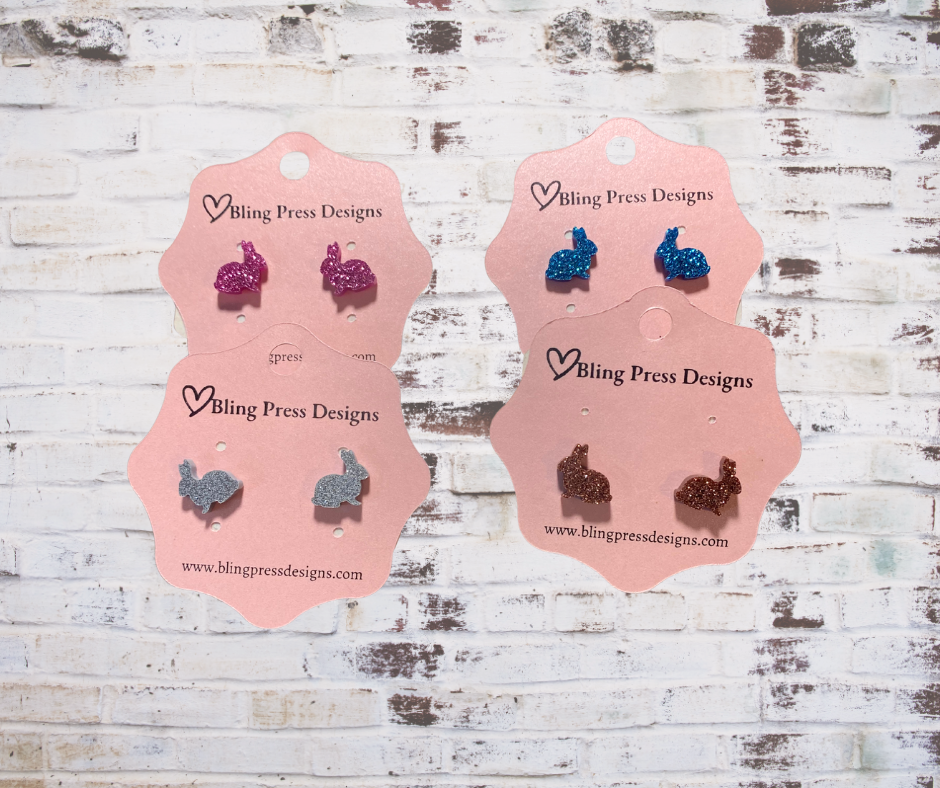 Four pairs of glitter bunny earrings on Bling Press Designs cards against a wooden background