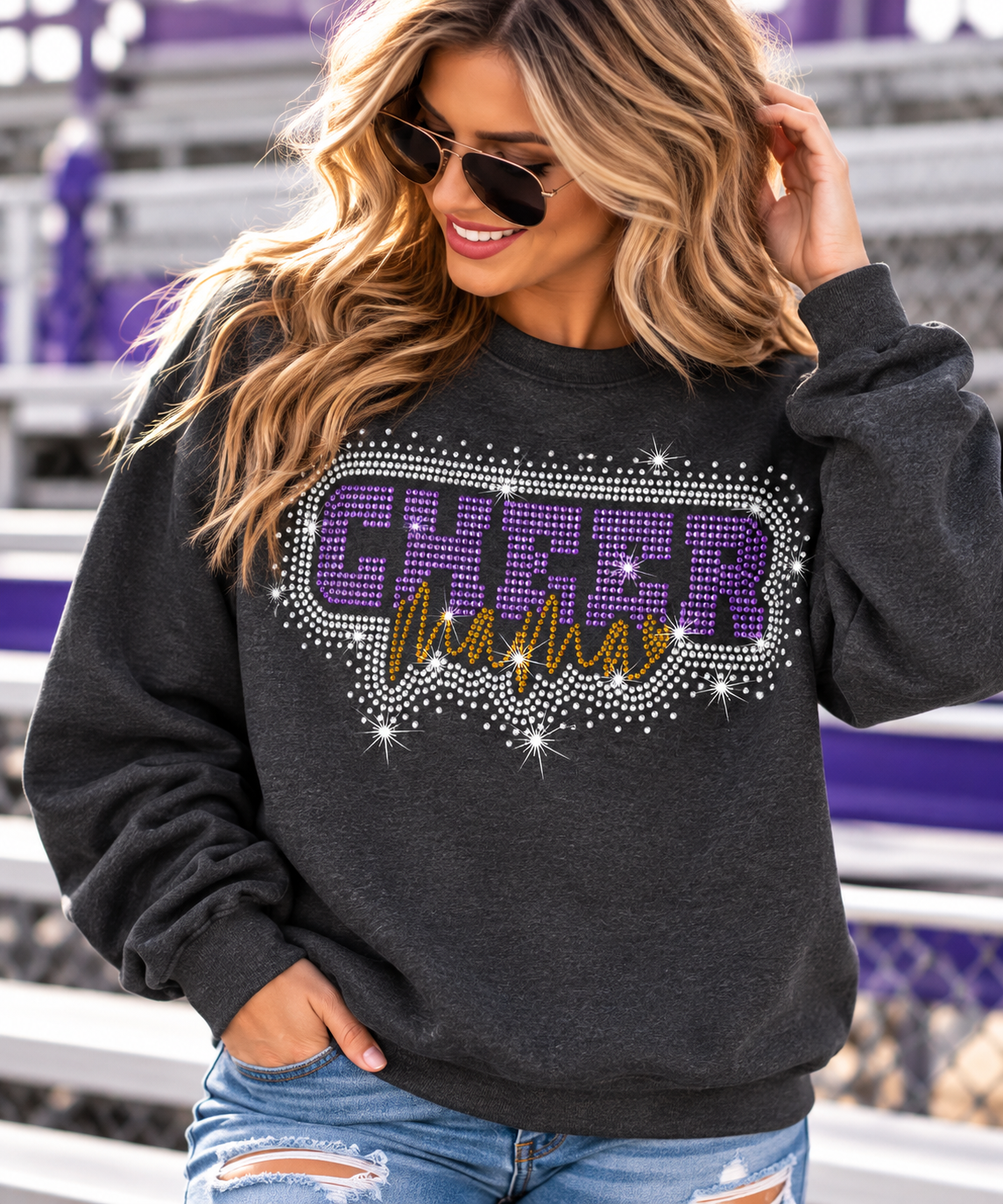 A woman wearing a dark charcoal gray sweatshirt with a rhinestone-embellished 'CHEER Mama' graphic in purple and gold, paired with distressed blue jeans, photographed outdoors in front of purple and white stadium seating.