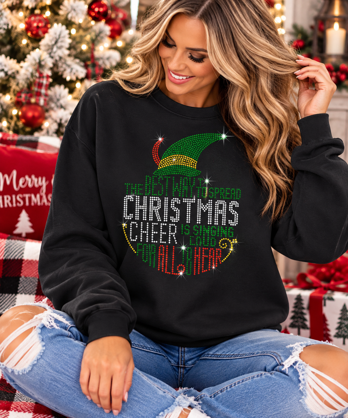 A woman wearing a black Christmas-themed sweatshirt with a festive green elf hat and rhinestone text that reads 'The Best Way to Spread Christmas Cheer is Singing for All Around,' posed in a decorated holiday setting.