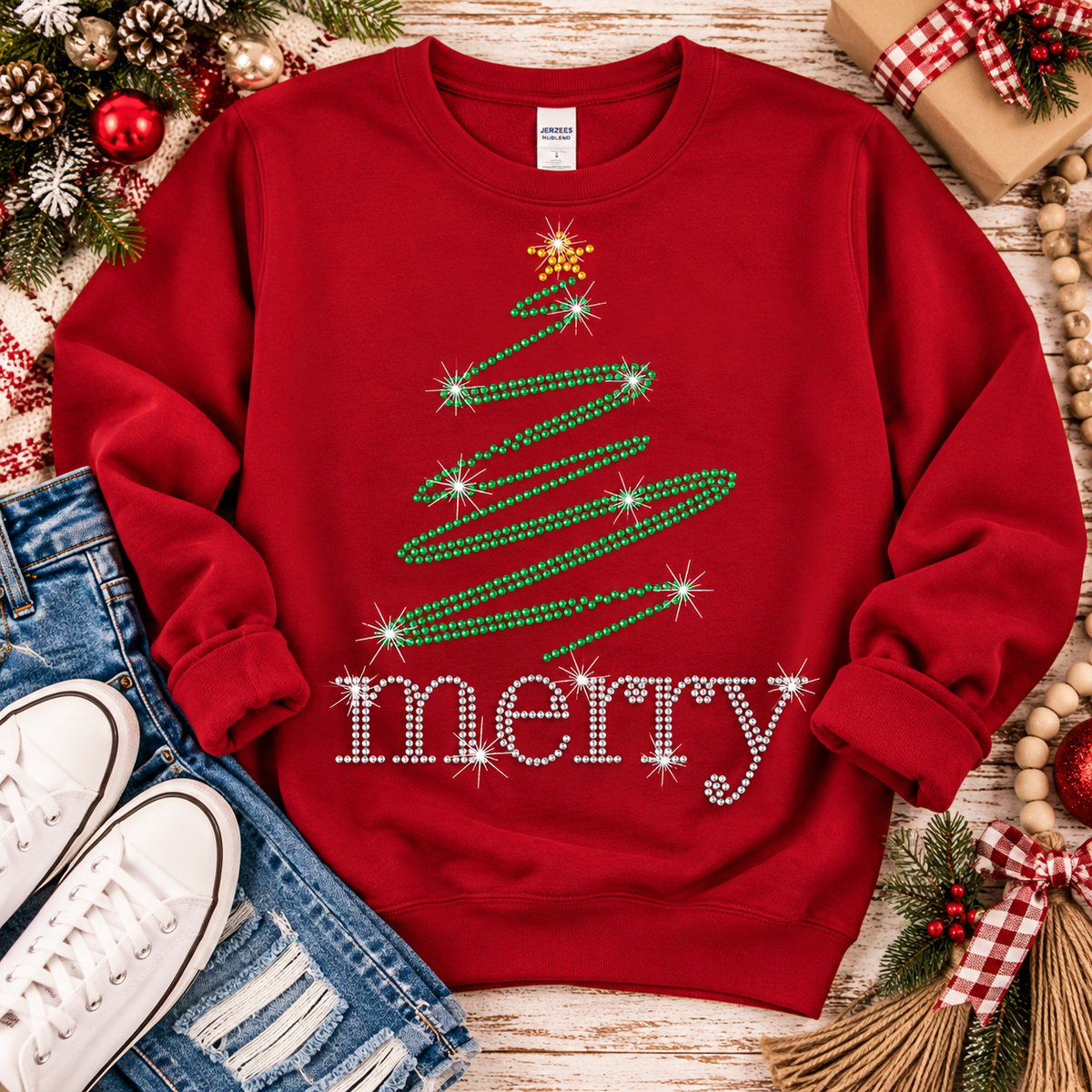 A red crewneck sweatshirt with a rhinestone Christmas tree and 'merry' text graphic, styled in a flat lay with jeans, sneakers, and holiday decorations.
