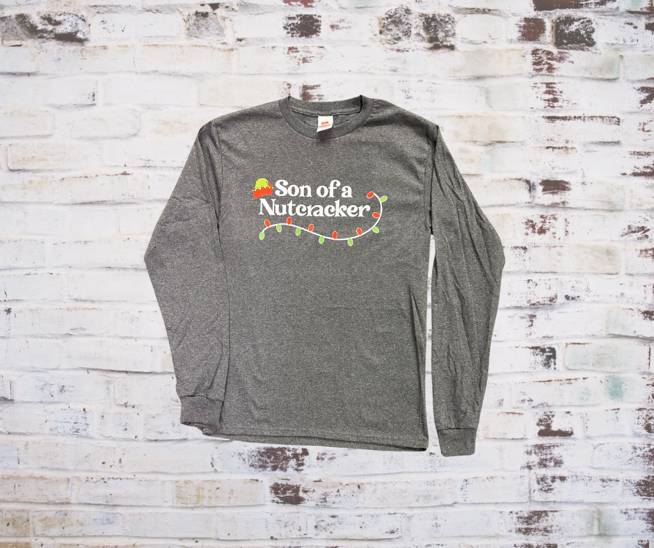 A **Son of a Nutcracker Long Sleeve Tee** featuring the playful phrase "Son of a Nutcracker" in bold lettering. Made from soft, breathable fabric for a comfortable, festive look during the holiday season.