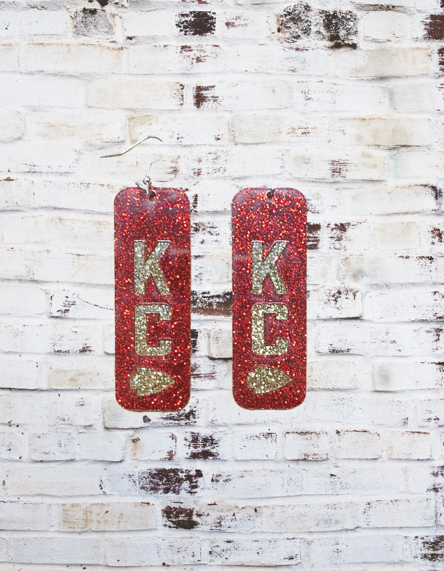 A pair of 2" glitter acrylic dangle earrings featuring a bold "KC" design, perfect for Kansas City fans, with a sparkling finish and lightweight construction for all-day wear.