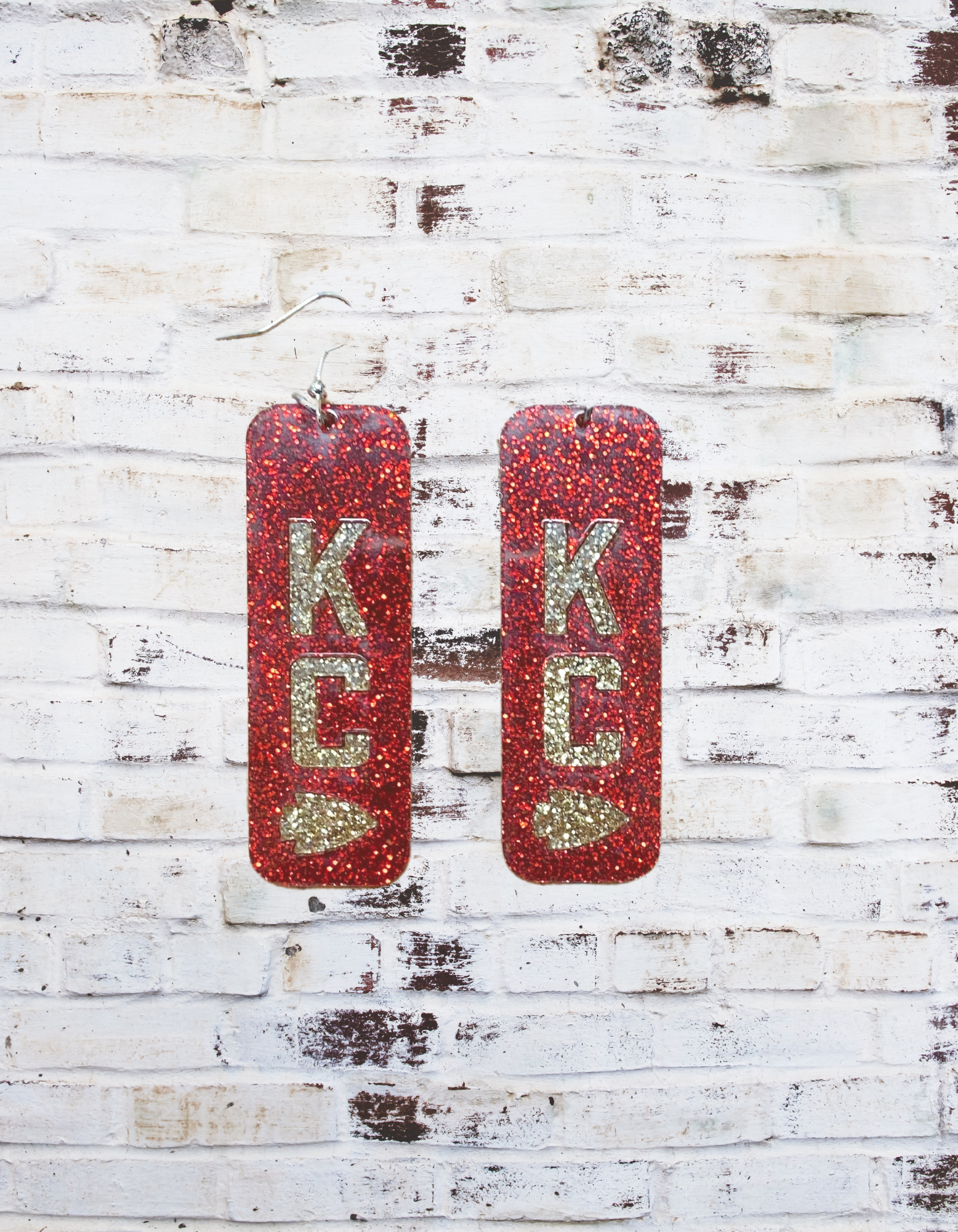 A pair of 2" glitter acrylic dangle earrings featuring a bold "KC" design, perfect for Kansas City fans, with a sparkling finish and lightweight construction for all-day wear.