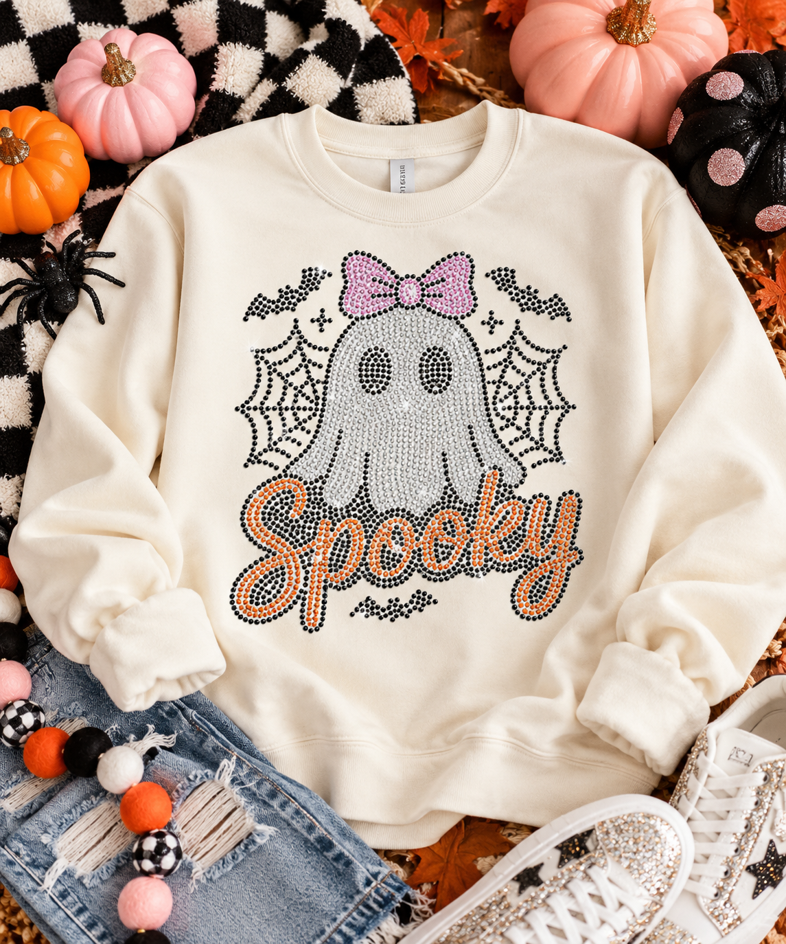 An off-white crewneck sweatshirt with a rhinestone-embellished ghost and 'Spooky' graphic, styled in a flat lay with pumpkins, jeans, sneakers, and autumn decorations.