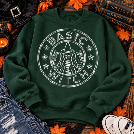 A dark green crewneck sweatshirt with a large white rhinestone-style graphic reading 'BASIC WITCH' around a witch illustration, styled in an autumn flat lay with books, fall leaves, a candle, jeans, and sneakers.
