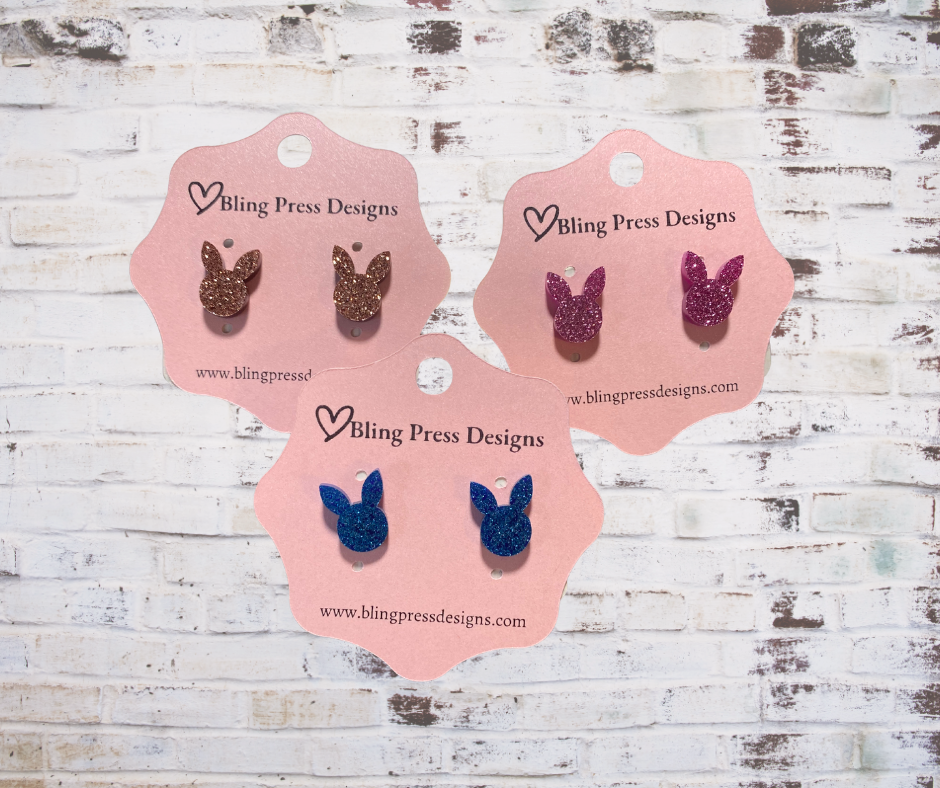 Three pairs of bunny ears-shaped earrings on Bling Press Designs cards against a wooden background.
