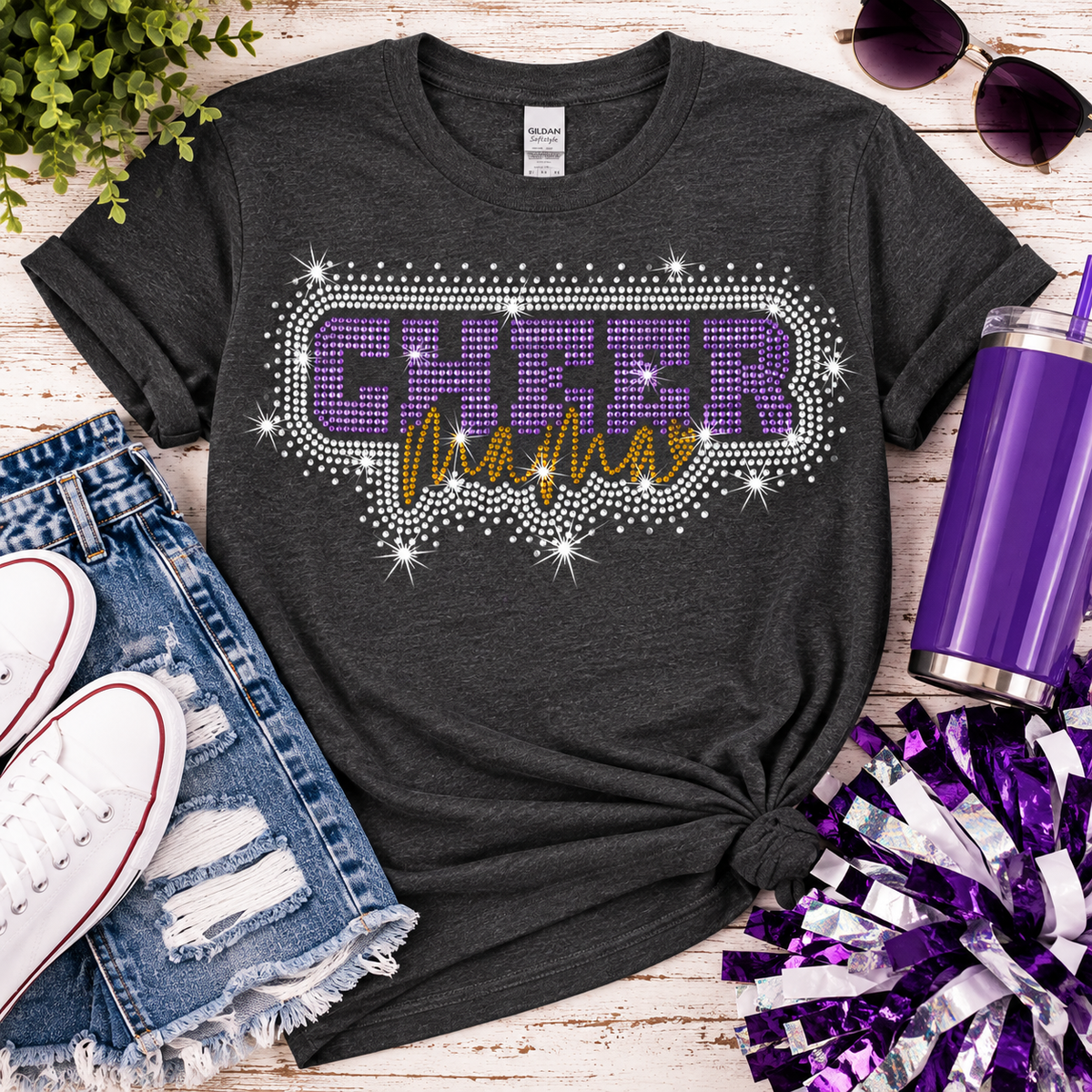 A dark charcoal gray graphic t-shirt with 'CHEER MAMA' written in purple and gold rhinestone-style lettering, styled in a flat lay with denim shorts, white sneakers, sunglasses, a purple tumbler, and purple pom-poms on a whitewashed wood background.