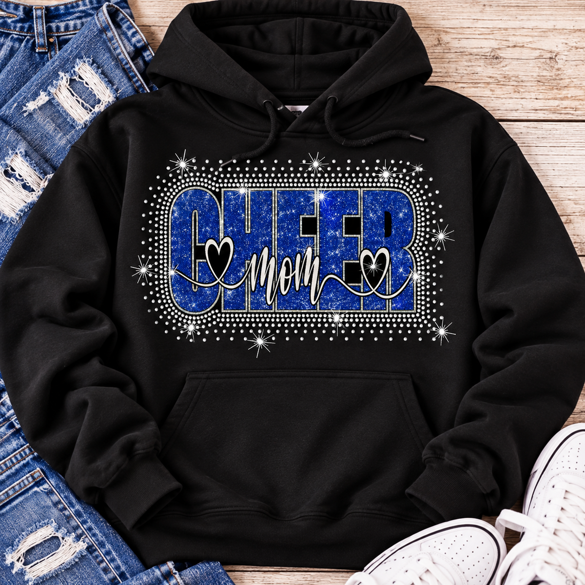 A black pullover hoodie laid flat on a wooden surface, styled with distressed blue jeans, white sneakers, and a rhinestone-style chest graphic that reads "CHEER mom" in blue and white with heart details.