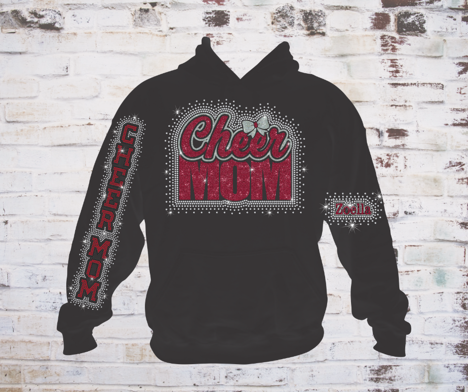 Black hoodie with a glitter and rhinestone design that reads ‘Cheer Mom’ accented with a bow with a rhinestone scatter around it,  the words "cheer mom in rhinestones on the sleeve, and athlete's name in glitter surrounded in rhinestones on the other sleeve