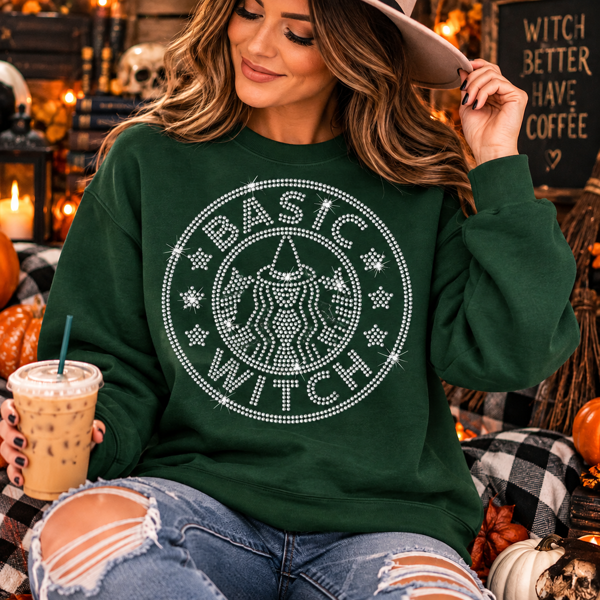 A woman wearing a dark green crewneck sweatshirt with a large white rhinestone graphic that reads 'BASIC WITCH' in a circular design. She is sitting in a cozy, autumn-themed setting with pumpkins, a coffee cup, and a hat, holding an iced coffee.