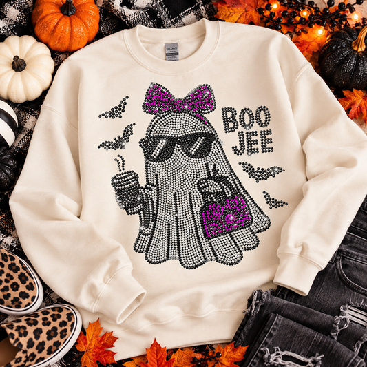 An off-white crewneck sweatshirt with a rhinestone-embellished ghost graphic wearing sunglasses and a purple bow, next to the text 'BOO JEE'. The sweatshirt is styled in a flat lay with autumn props including pumpkins, leaves, and denim.