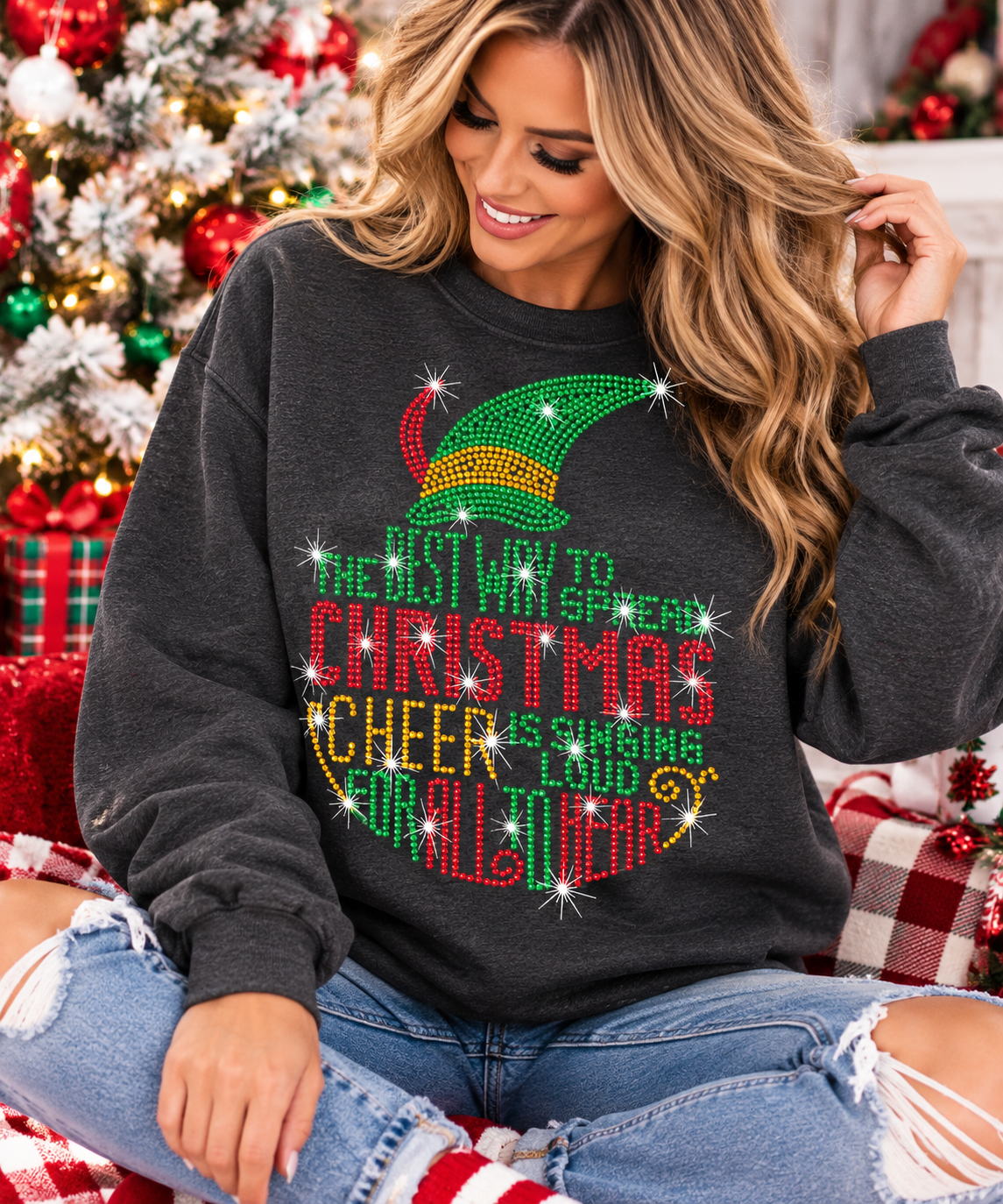 A woman wearing a dark gray Christmas-themed sweatshirt with a large rhinestone elf hat and colorful text reading 'The Best Way to Christmas is Cheering All Year Long.' She is seated in a decorated holiday setting with a Christmas tree and plaid blankets in the background.