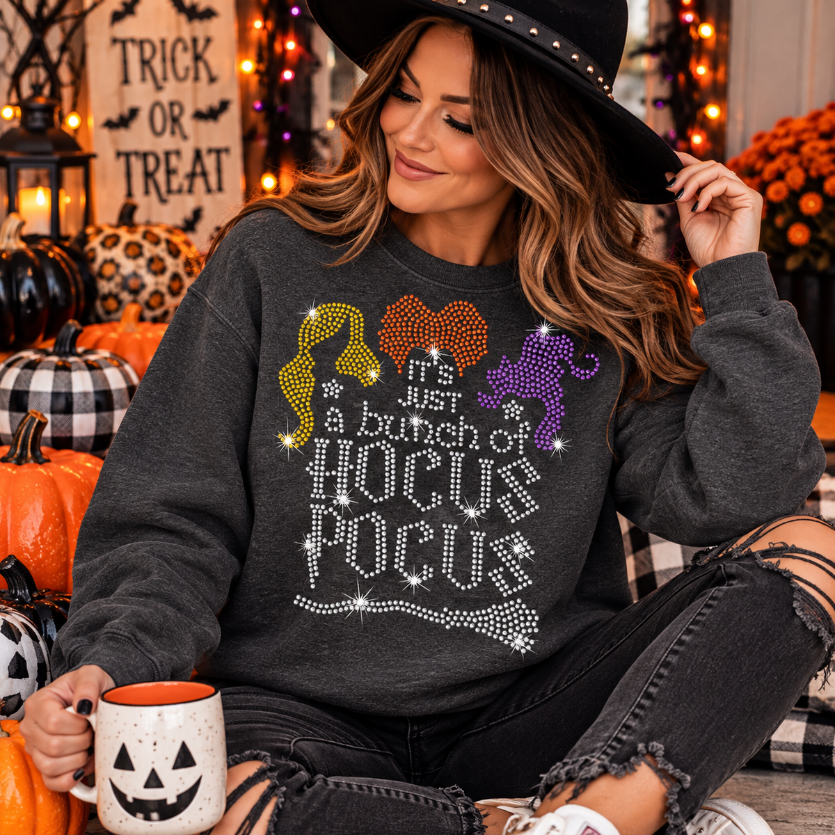 A woman wearing a dark gray crewneck sweatshirt with a rhinestone 'Hocus Pocus' graphic sits in a decorated Halloween setting with pumpkins, a mug, and a sign that reads 'Trick or Treat'.