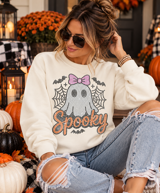 A woman wearing a cream-colored crewneck sweatshirt with a 'Spooky' ghost graphic, paired with ripped light-wash jeans, posed in a festive autumn setting with pumpkins and candles.