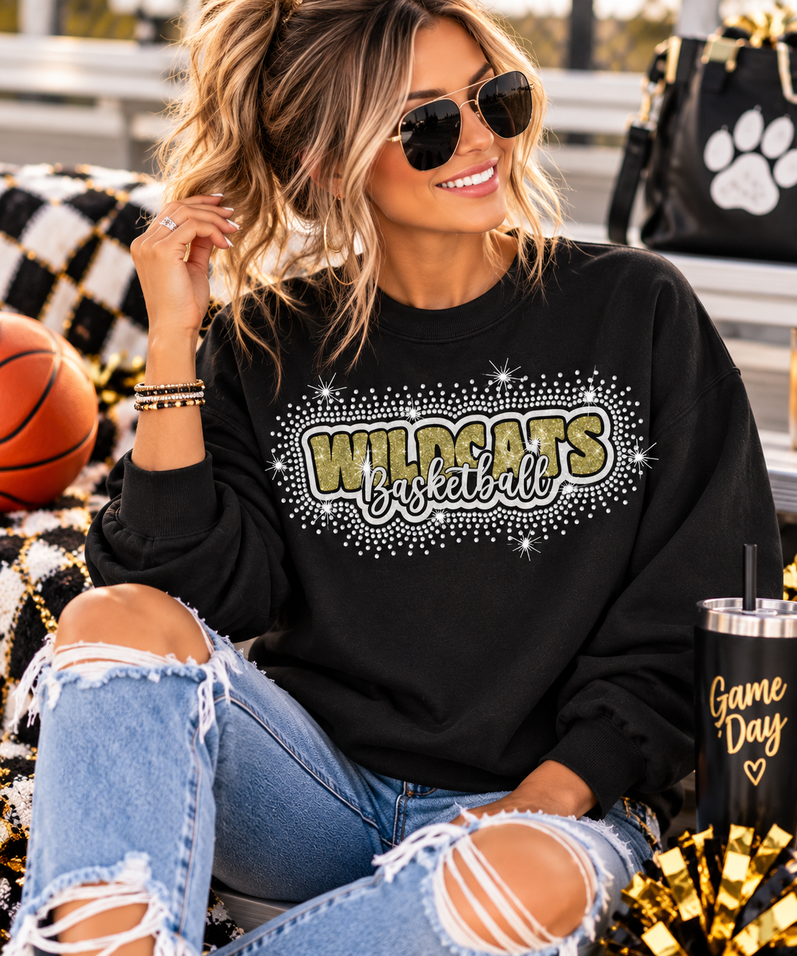 A woman wearing a black crewneck sweatshirt with a 'Wildcats Basketball' graphic printed across the chest in gold and white with a rhinestone border. She is also wearing ripped blue jeans and sunglasses, sitting in a decorated outdoor setting with a basketball and a black tumbler nearby.