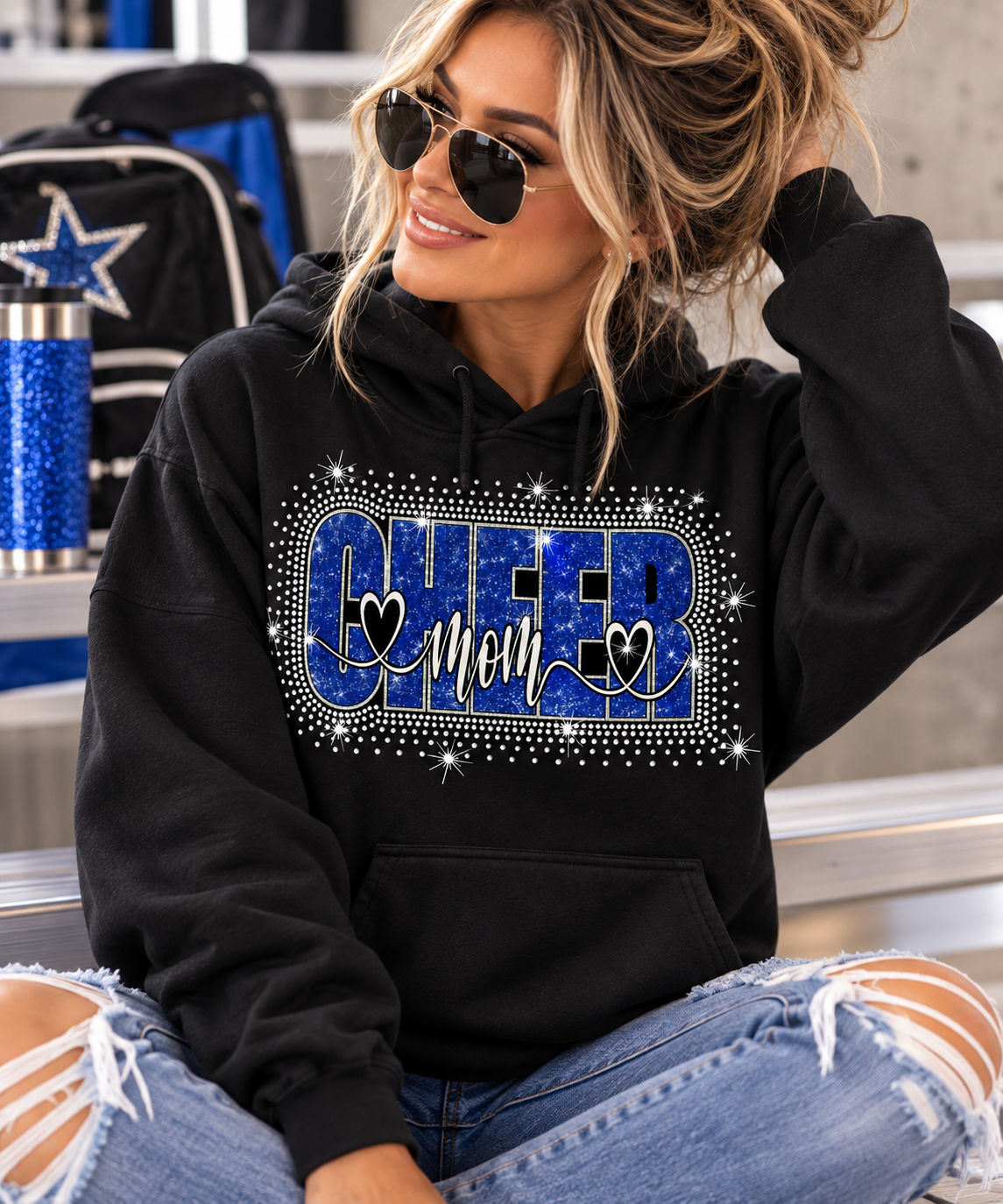 A woman wearing a black hoodie with a blue and silver rhinestone-style 'CHEER mom' graphic, seated in a locker room setting.