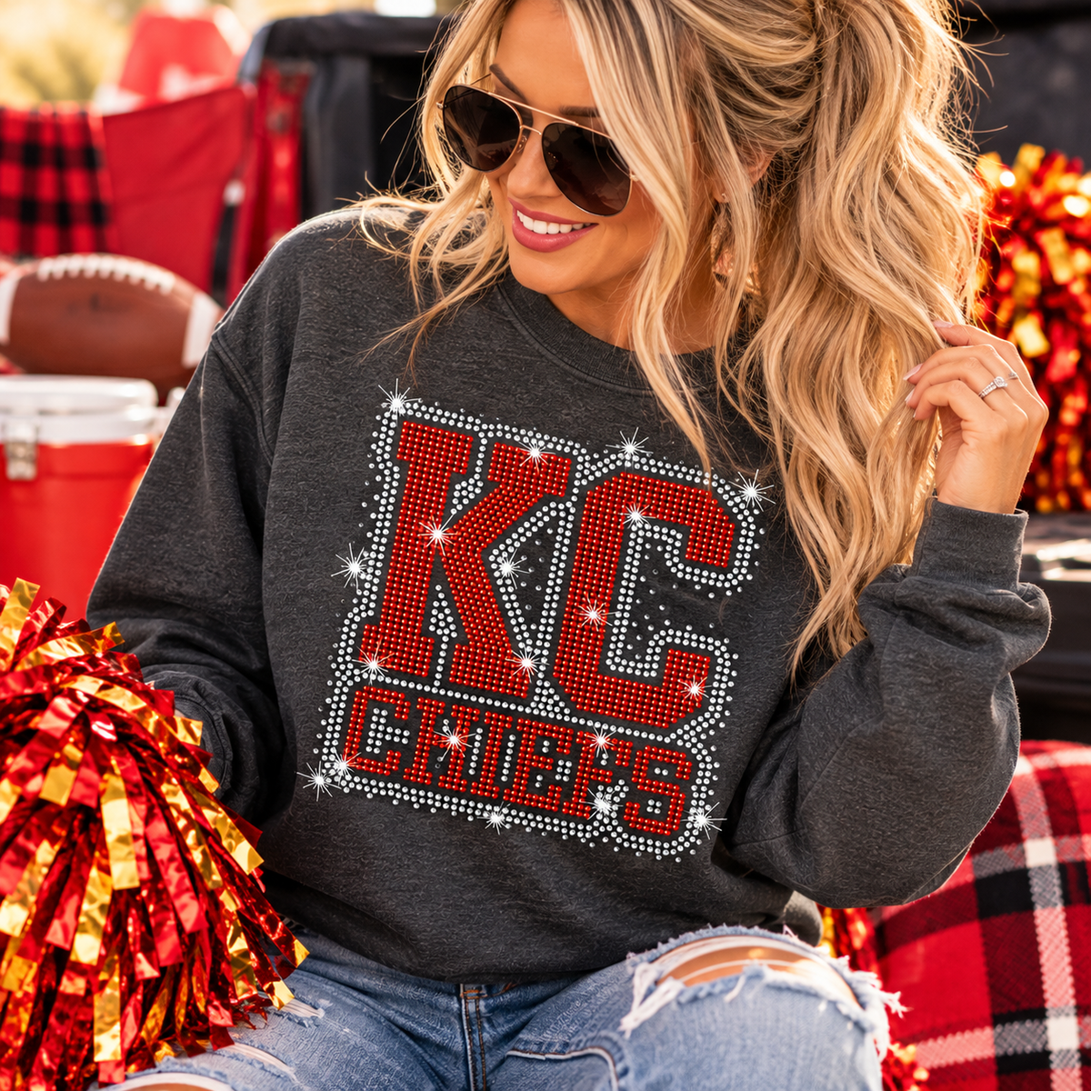 A woman wearing a dark gray crewneck sweatshirt with a large red and silver rhinestone 'KC CHIEFS' graphic on the front, paired with ripped blue jeans and sunglasses, holding red and gold pom-poms in a festive outdoor setting.