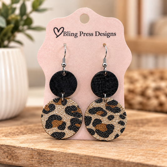 A pair of round drop earrings displayed on a pink card. Each earring has a small black round top and a larger glittery leopard-print circle below in beige, brown, and black.