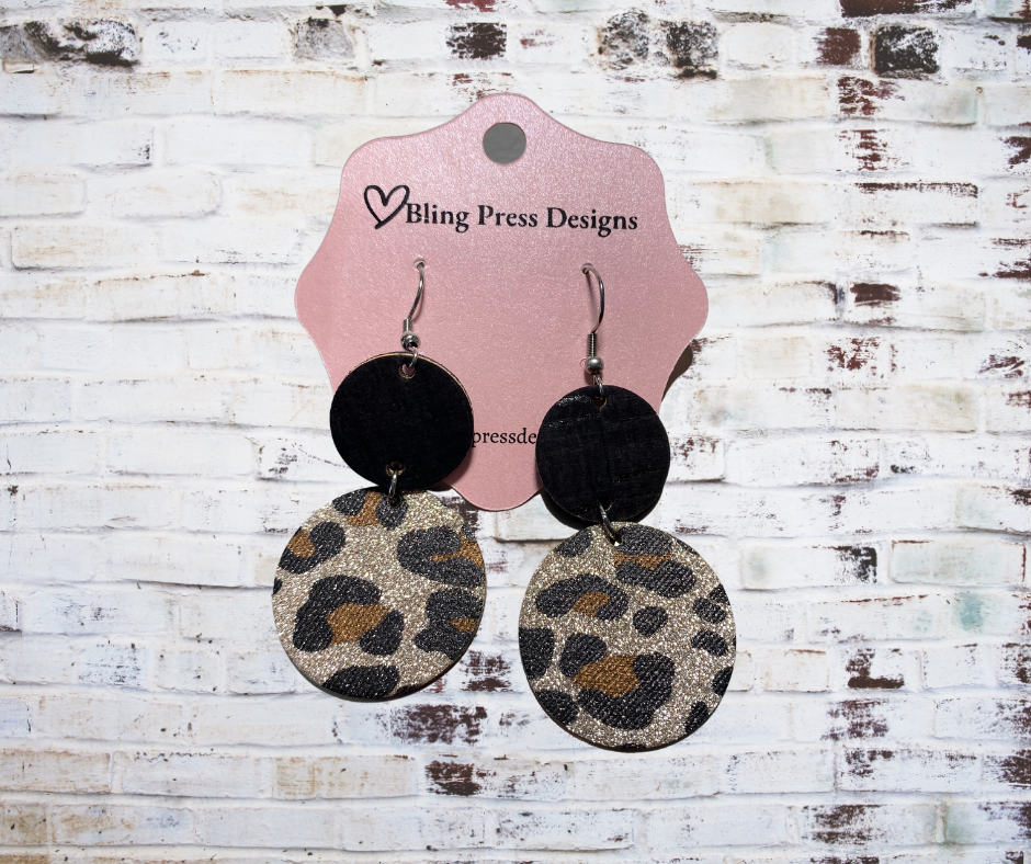 A pair of bold corky glitter stacked leopard dangle earrings, featuring lightweight cork with a glitter leopard print, black accents, and layered geometric shapes, hanging from hypoallergenic hooks. Perfect for adding sparkle and style to any outfit.