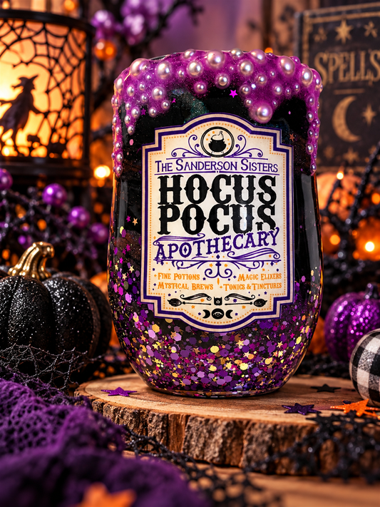 A Halloween-themed decorative bottle labeled 'The Sanderson Sisters Hocus Pocus Apothecary' filled with dark liquid and covered in purple beads and gold glitter, set on a wood slice with witch lanterns and purple fabric in a styled scene.