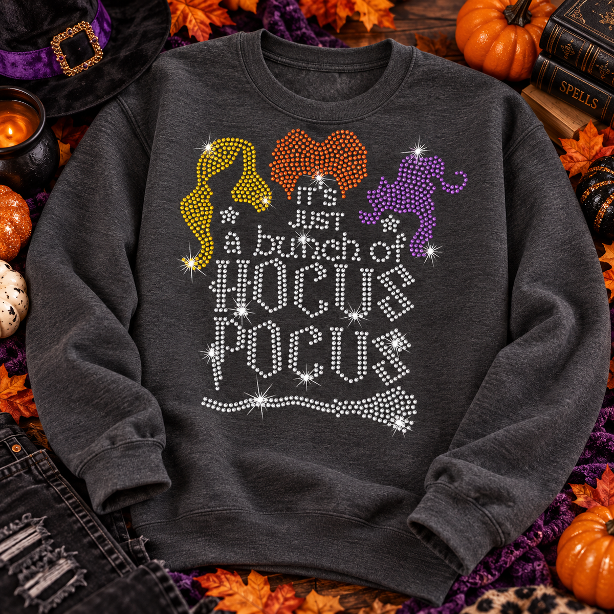 A dark gray crewneck sweatshirt with a rhinestone 'Hocus Pocus' graphic sits in a decorated Halloween setting with pumpkins, a mug, and a sign that reads 'Trick or Treat'