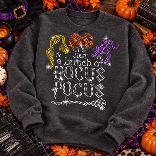 A dark gray crewneck sweatshirt with a rhinestone 'Hocus Pocus' graphic sits in a decorated Halloween setting with pumpkins, a mug, and a sign that reads 'Trick or Treat'