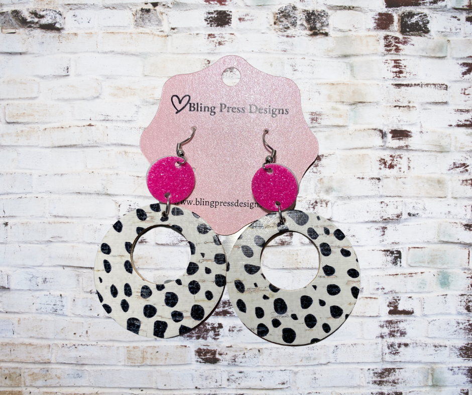 Earrings with pink circular tops and black and white polka dot designs on a wooden background with Bling Press Designs card.