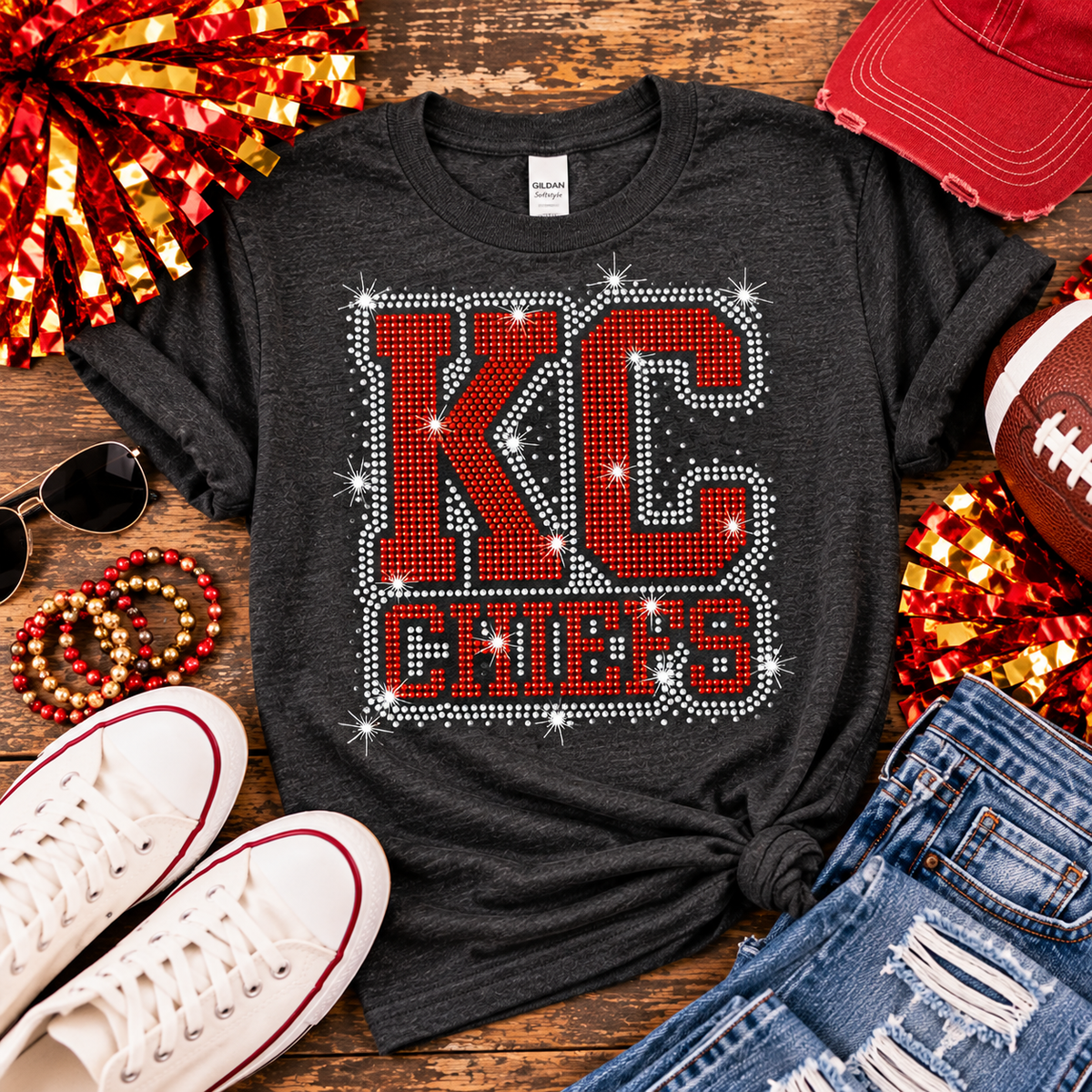 A dark charcoal gray graphic t-shirt featuring 'KC CHICKS' in red block letters outlined with white rhinestones, styled in a flat lay with white sneakers, blue jeans, a red baseball cap, sunglasses, beaded bracelets, and sports props on a wooden background.