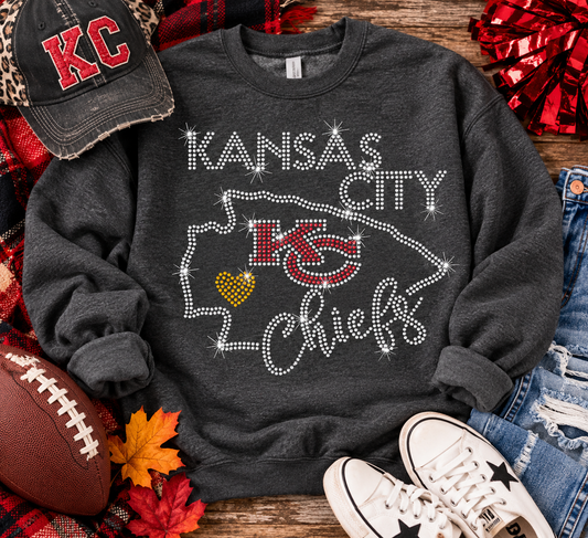 A dark charcoal gray sweatshirt with 'Kansas City Chiefs' text and a Chiefs logo outlined in rhinestones, styled in a flat lay with a baseball cap, football, jeans, sneakers, and autumn leaves on a wooden surface.
