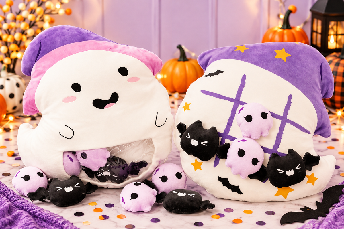 Two plush ghost-shaped pillows with Halloween-themed designs, including one with a smiling face and one with a tic-tac-toe grid and black cat faces, displayed with small plush bats and pumpkins on a festive table setting.