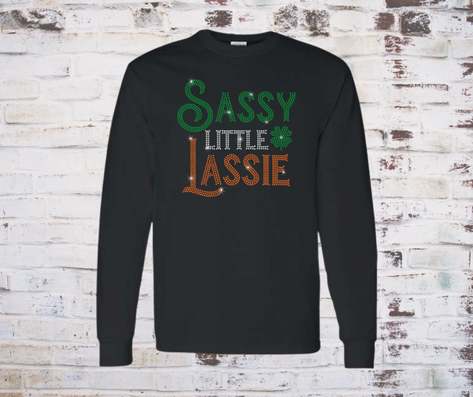 Black long-sleeve shirt with 'Sassy Little Lassie' rhinestone text on a wooden background