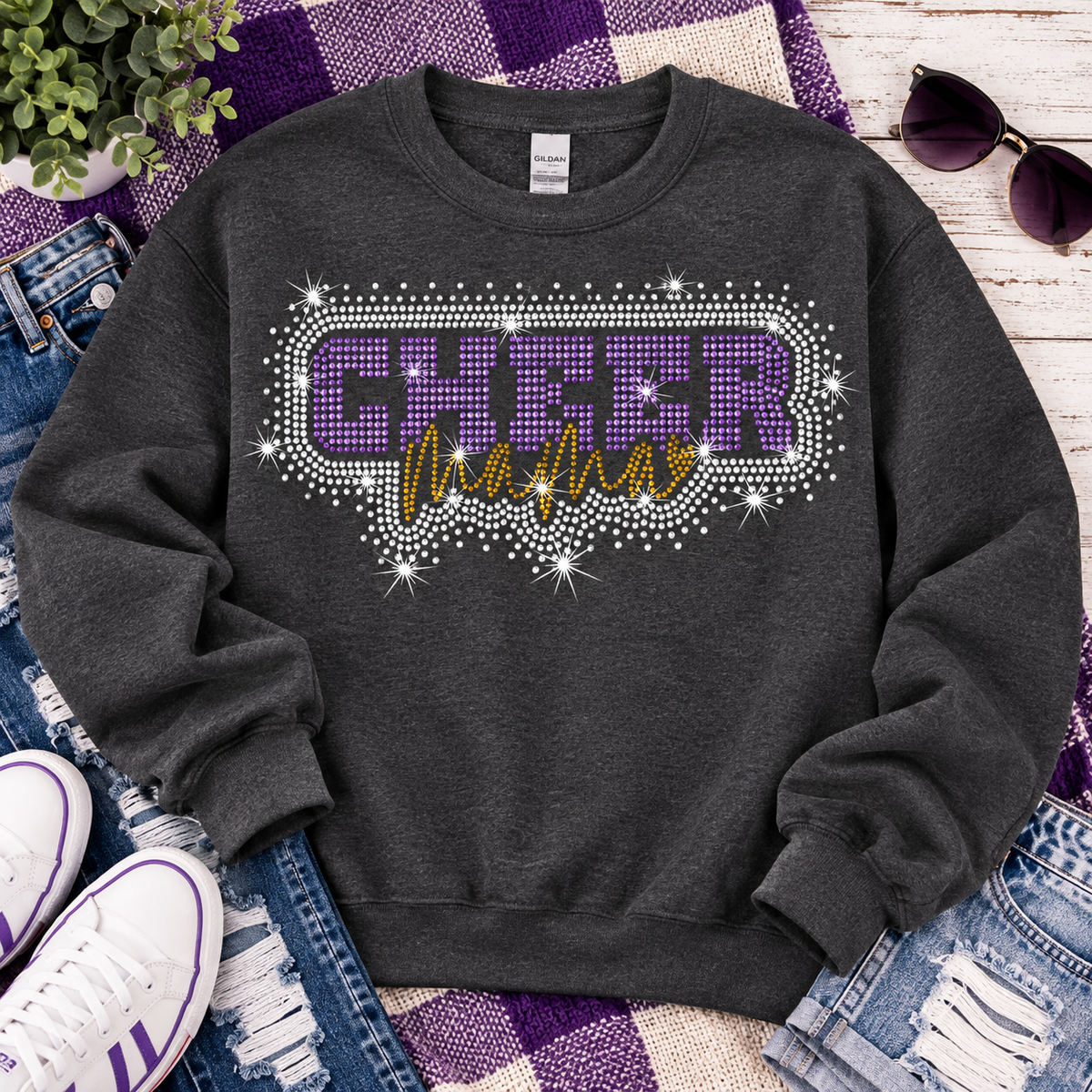 A dark heather gray crewneck sweatshirt with a rhinestone-embellished 'CHEER mama' graphic in purple and gold, styled in a flat lay with jeans, white sneakers, sunglasses, and a purple plaid blanket.
