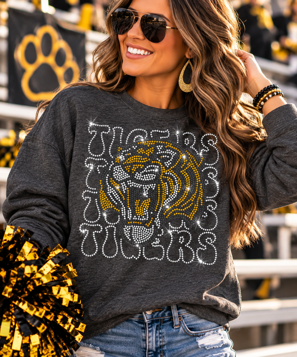 A woman wearing a dark gray long-sleeve sweatshirt with a large rhinestone lion graphic and the word 'TIGERS' in white and gold. She is in a stadium setting with gold and black pom-poms.