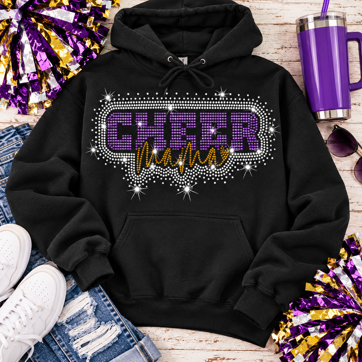 A black pullover hoodie laid flat on a rustic wood surface, featuring a large rhinestone-style chest graphic that reads 'CHEER Mamas' in purple and gold. The hoodie is styled with white sneakers, blue jeans, a purple tumbler, sunglasses, and purple-and-gold pom-poms.