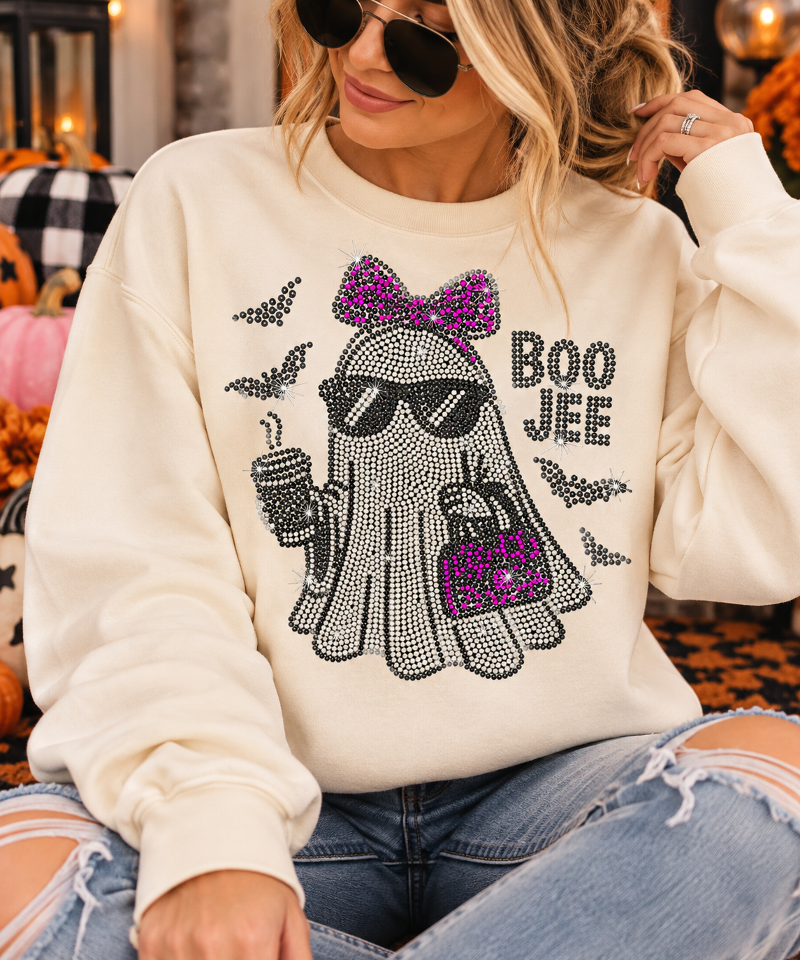 A woman wearing a cream-colored sweatshirt with a rhinestone ghost graphic that says 'BOO JEE' in black. The sweatshirt features a rhinestone ghost wearing sunglasses, a purple bow, and holding a drink and a purse, with black bat silhouettes.