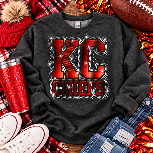A dark charcoal gray sweatshirt with a large 'KC CHIEFS' graphic made of red letters and white rhinestone-like sparkles, styled in a flat lay with a red pom-pom beanie, red tumbler, football, jeans, sneakers, and plaid fabric.