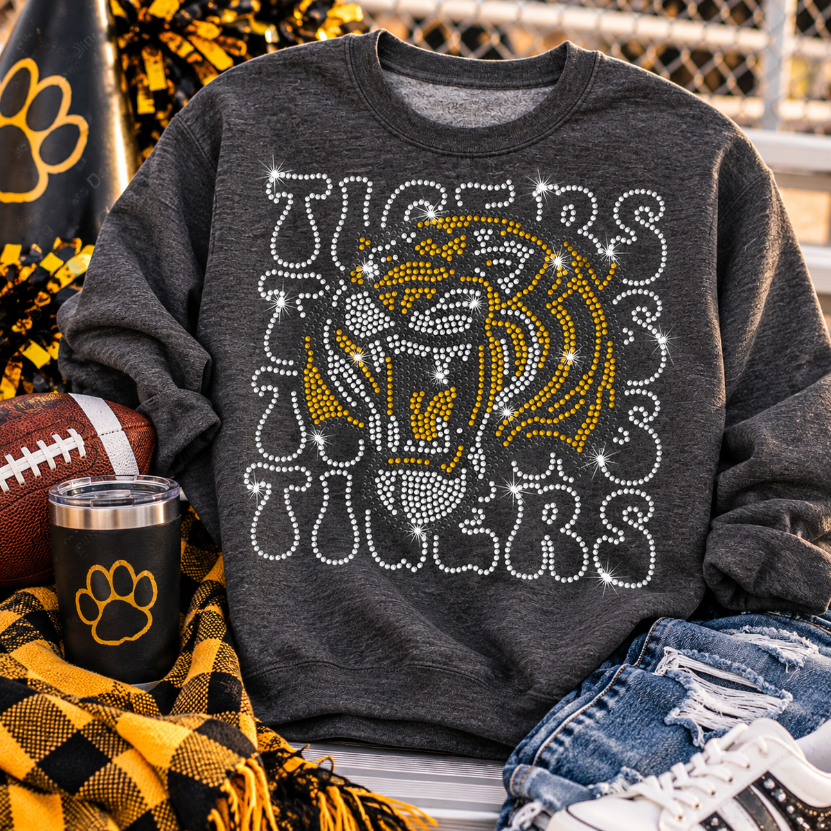 A dark heather gray sweatshirt with a large rhinestone tiger head and 'Tigers' text graphic, styled in a football-themed flat lay with a plaid blanket, tumbler, football, jeans, and sneakers.