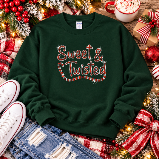 A dark green crewneck sweatshirt with 'Sweet & Twisted' written in red and white candy cane lettering, styled in a festive flat-lay with jeans, white sneakers, a mug of hot cocoa, and Christmas decorations.