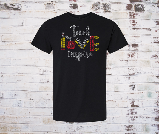 Black t-shirt with 'Teach Love Inspire' rhinestone text and graphics on a wooden background