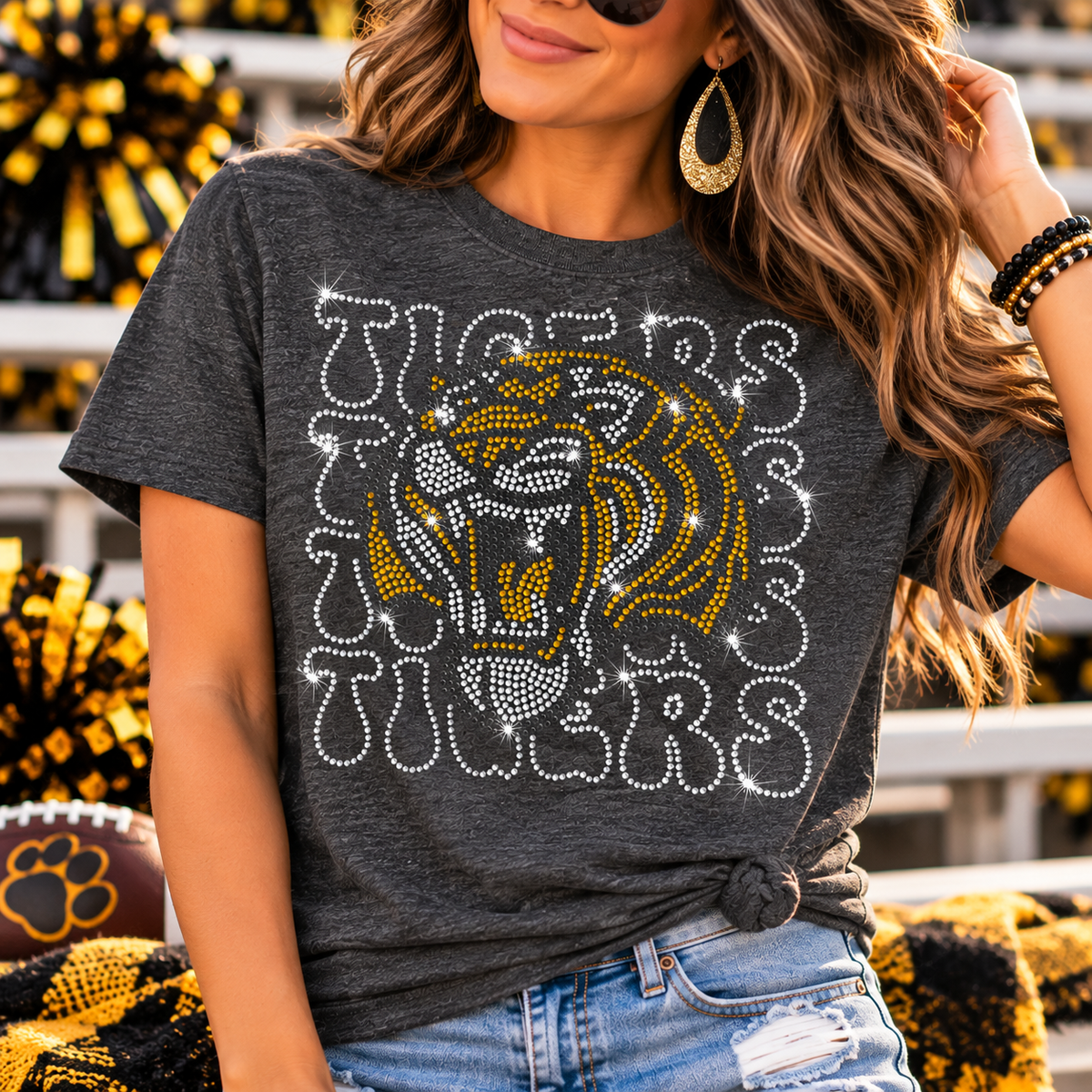 A woman wearing a dark heather gray graphic t-shirt with a large rhinestone tiger head and 'TIGERS' text in white and gold, paired with blue jeans.