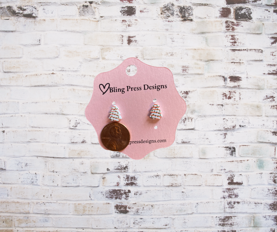 Small Christmas tree cake stud earrings shown next to a penny for size reference, featuring a festive green tree design with holiday details.
