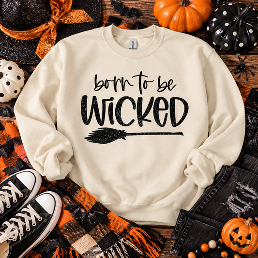 An off-white crewneck sweatshirt with 'born to be Wicked' and a broom graphic printed in black, styled in a flat lay with Halloween props including pumpkins, a plaid scarf, jeans, and sneakers.