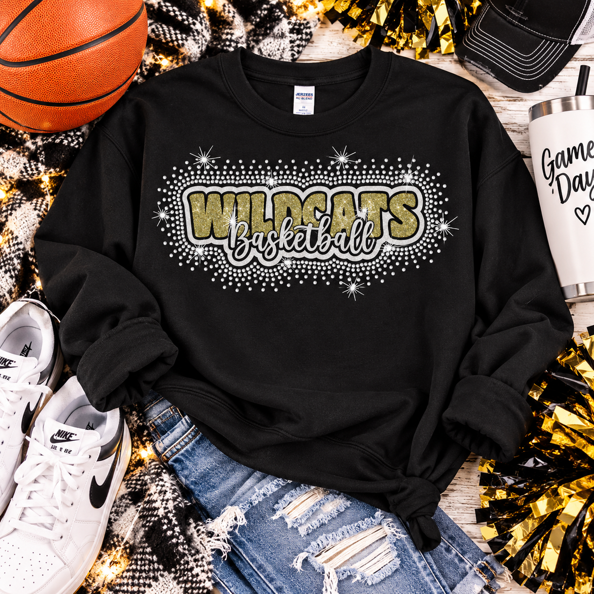 A black long-sleeve sweatshirt laid flat with rolled cuffs, featuring a large 'Wildcats Basketball' graphic across the chest in glittery gold and silver. The sweatshirt is styled in a flat lay with a basketball, white sneakers, ripped blue jeans, a tumbler, and gold tinsel on a wood surface.
